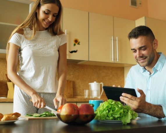 Attractive couple chatting in the kitchen early morning. Handsome man using tablet while his girlfriend cooking breakfast at home